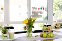 Im Vordergrund ein dekorierter Tisch mit gelben Blumen, Obst, einer Platte mit Kerze und Pflanze. Im Hintergrund weitere geschmückte Tische und eine helle Fensterfront.